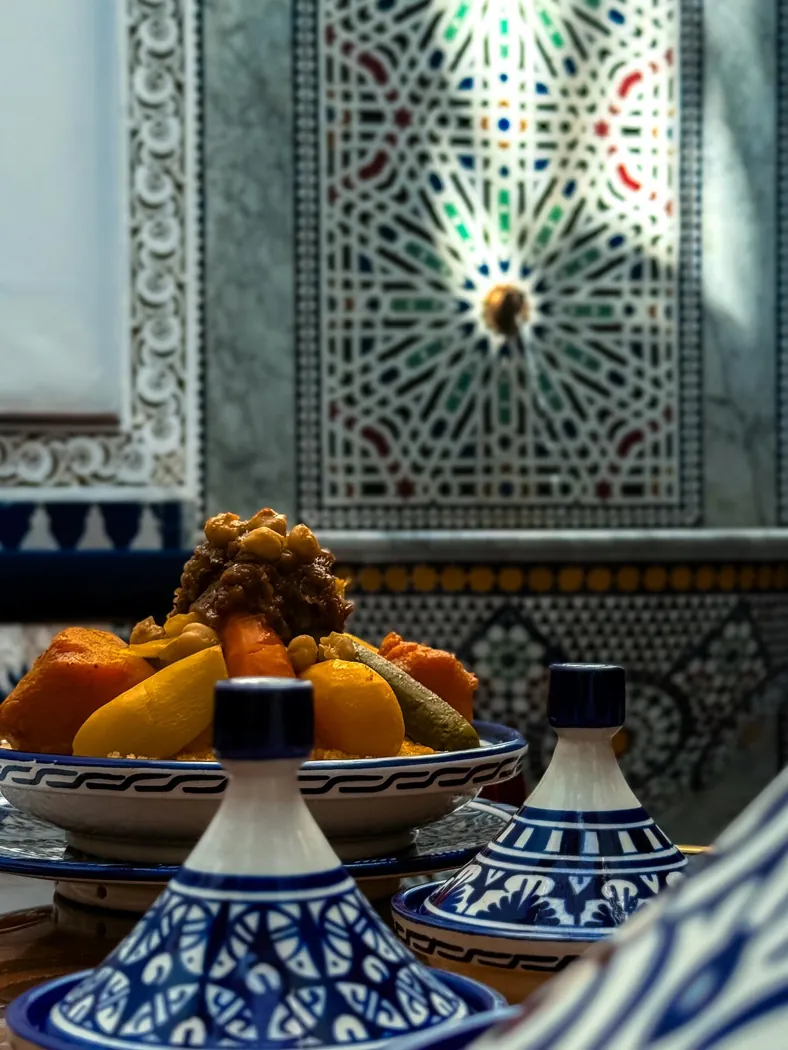 Tajines en céramique bleue et coupe de fruits devant une fontaine en zellige chez MaQam