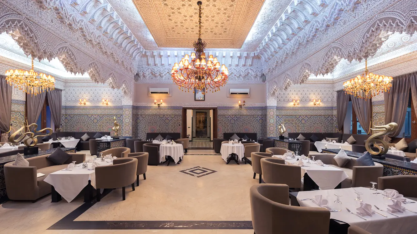 Grande salle de Riad 21 à Casablanca : plafond en plâtre ciselé, lustres et tables nappées de blanc
