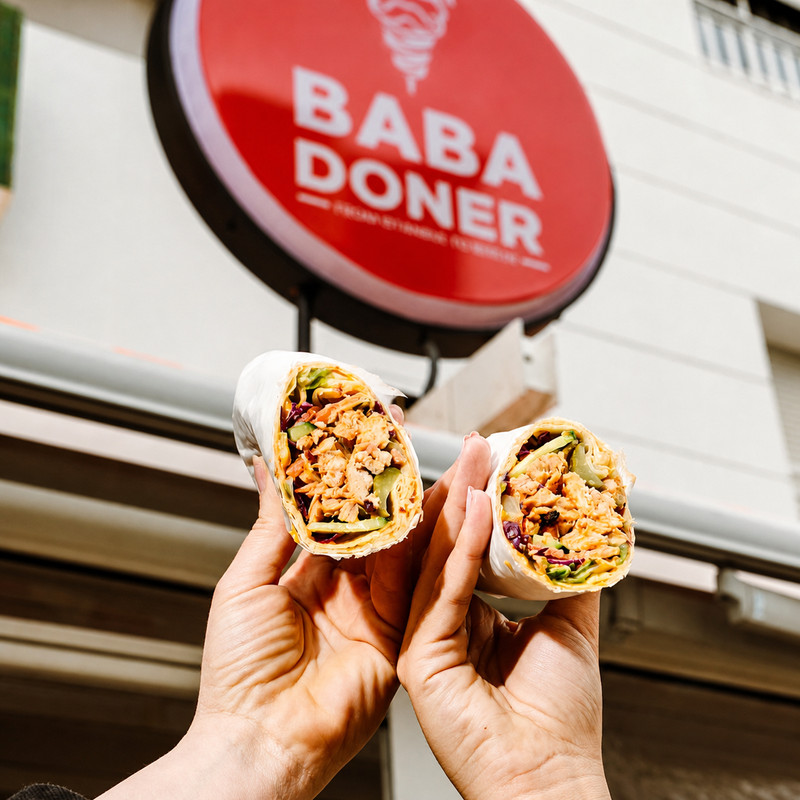 Deux döners roulés tenus devant l'enseigne ronde rouge Baba Doner