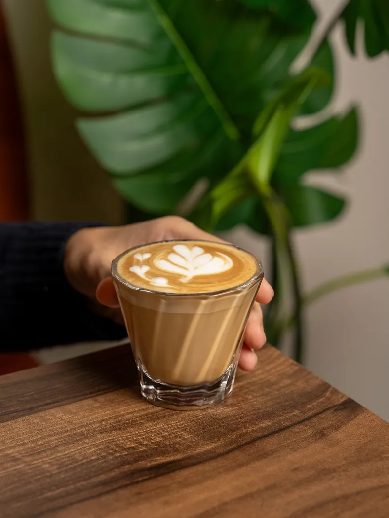 Cappuccino avec dessin de latte art tenu à la main, plante verte en fond