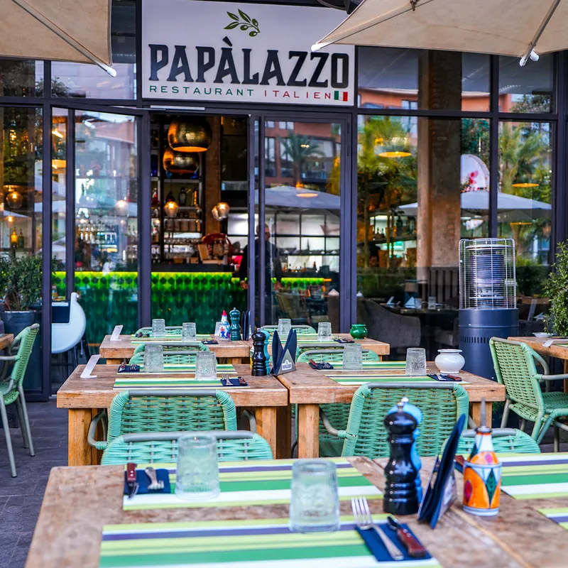 Papàlazzo à Marrakech : terrasse de M Avenue, tables en bois et chaises vertes devant l'enseigne