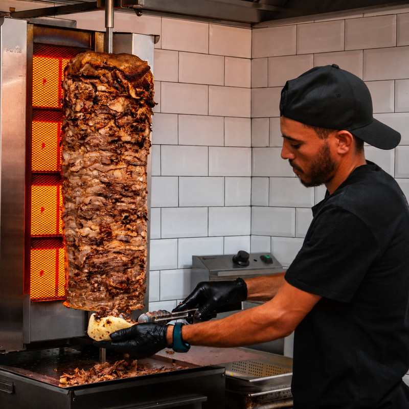 Cuisinier de Kakou Kebab tranchant la broche de kebab devant un mur carrelé blanc
