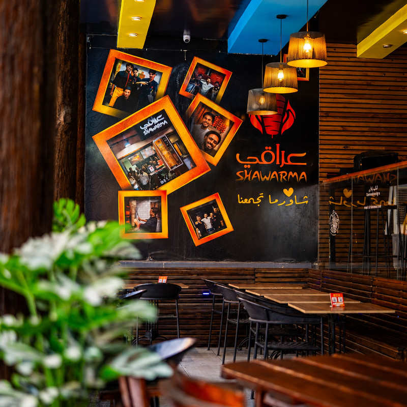 Salle d'Iraki Shawarma, mur noir au logo orange et cadres photo, suspensions en osier